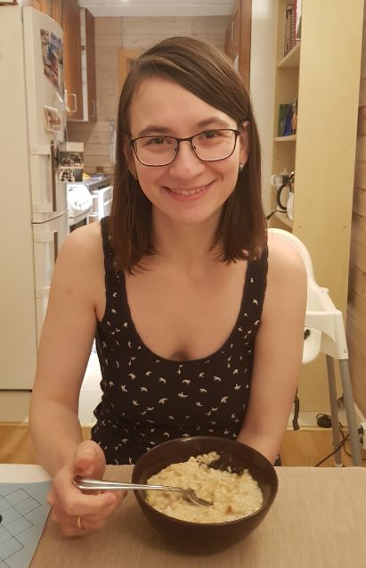 A picture of a woman with long hair, in a tank top, sitting by a dining table, smiling at the camera with a bowl of oatmeal in front of her. Her arms are somewhat thin and there are slight bags under her eyes.