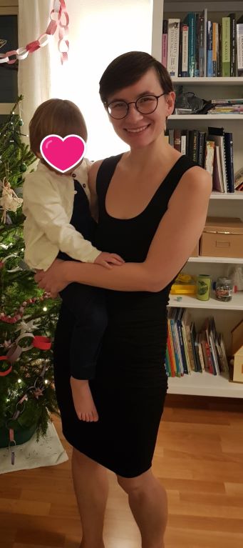 A picture of a woman in a black dress holding a child on her hip, standing by a Christmas tree.