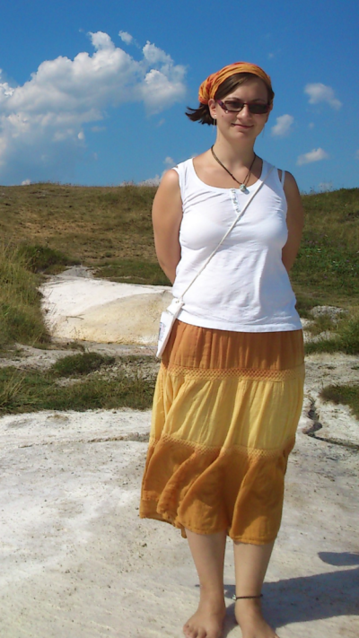 A picture of a young woman with a long skirt and a tank top standing on a hill outside. Her feet are pointing out and the pelvis is in a posterior tilt.